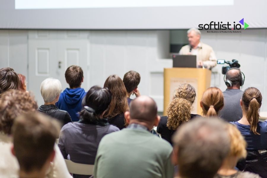 Audience attending a presentation by a speaker at a podium with a "softlist.io" logo in the background in a conference room.